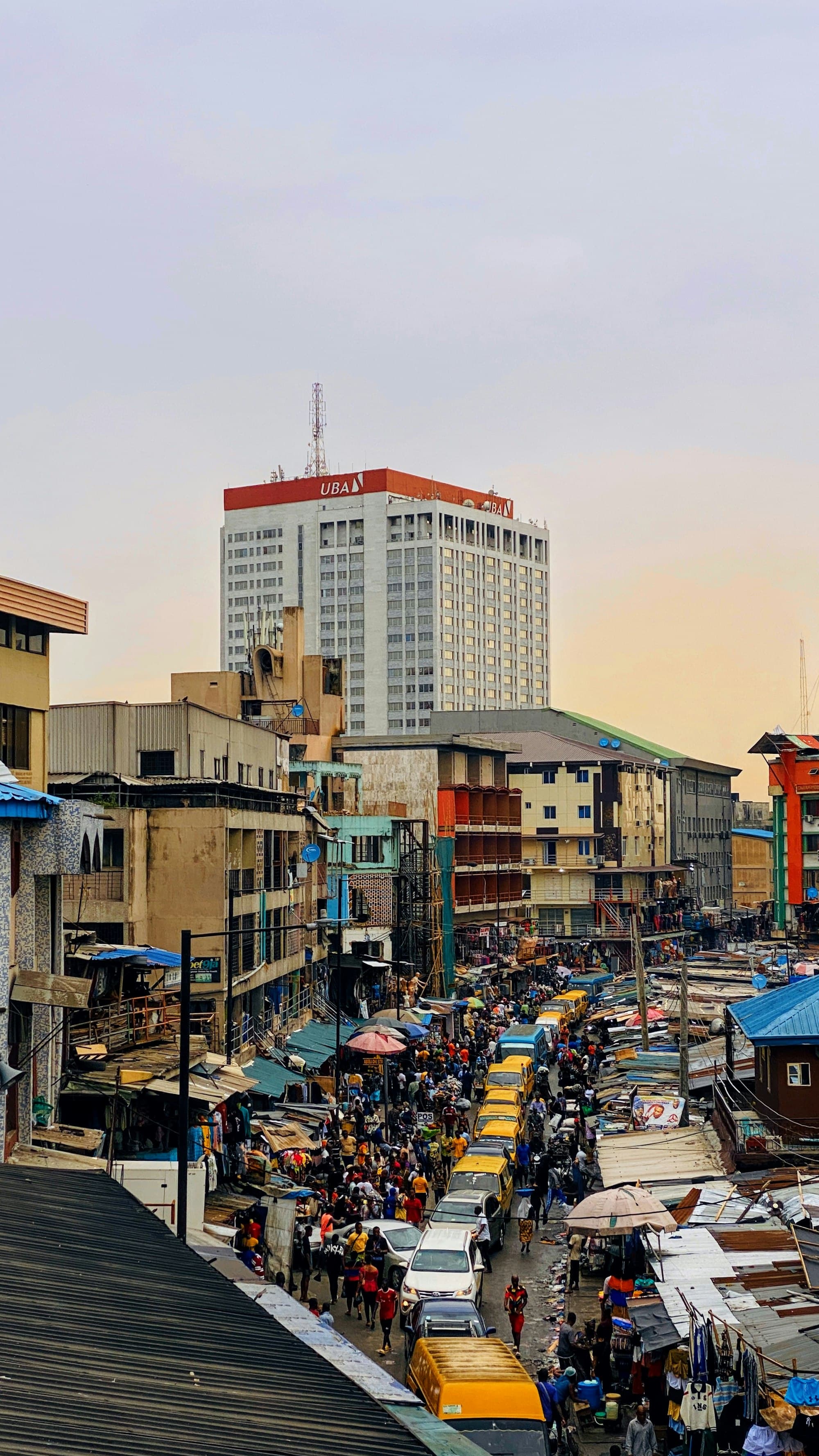 Lagos traffic at dusk, with yellow danfo buses in the street below a tall office tower